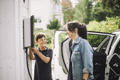 Smiling boy holding cable while looking at mother standing near electric car