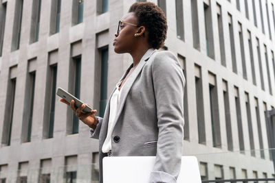 From below african american businesswoman in formal clothes standing near building with laptop while browsing on smartphone