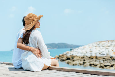 Rear view of couple sitting on pier by sea against sky