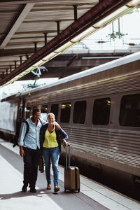 Full length of happy couple with luggage walking by train on railroad station platform