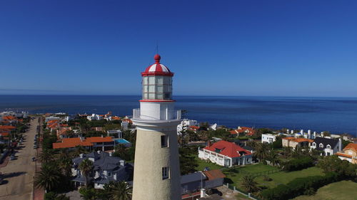 Lighthouse by sea against buildings in city against clear blue sky