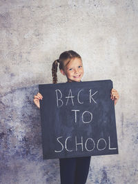 Portrait of smiling girl holding writing slate with text against wall