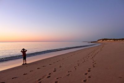 Rear view of boy standing at beach against clear sky during sunset