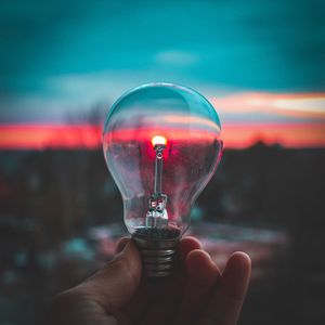 Close-up of hand holding light bulb against sky during sunset