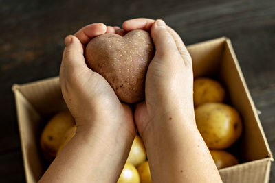 Female hands holding a heart-shaped ugly vegetable potato over a box filled with potatoes. 