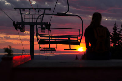 Silhouette woman sitting under ski lift against orange sky