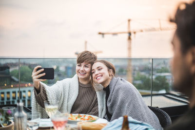 Smiling female friends taking selfie through phone with blanket while sitting on terrace during social gathering