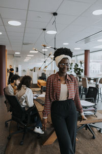 Portrait of smiling young businesswoman leaning on desk against colleagues discussing strategy in office