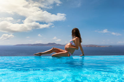 Pregnant woman sitting at infinity pool against sky