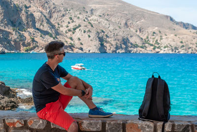 A man looks at the turquoise sea and mountains sitting on a stone parapet