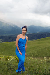 Woman in a blue long dress stands on top of a mountain in the summer on a green meadow