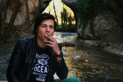 Portrait of young man smoking cigarette while sitting against stream