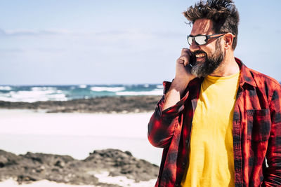 Man smiling while talking on mobile phone standing at beach against sky