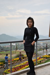 Portrait of young woman with hands in pockets standing against sky