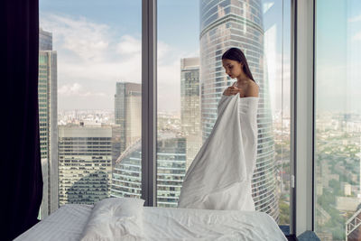 Young woman wrapped in a sheet stands in a bedroom in a skyscraper during the day