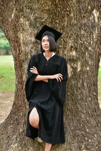 Young woman in graduation gown standing against tree