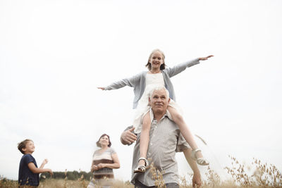 Happy girl sitting on her grandfather's shoulders in nature with brother and grandmother in the background