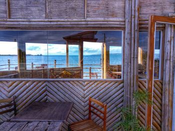 Wooden chairs by sea against sky seen through window