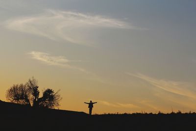 Silhouette man with arms outstretched standing on field against sky at sunset