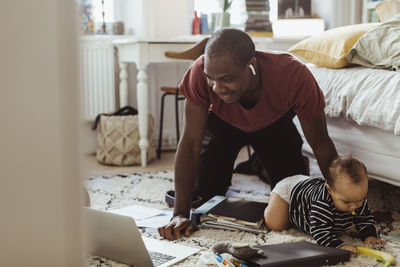 Smiling dadpreneur looking at laptop while kneeling behind son in bedroom