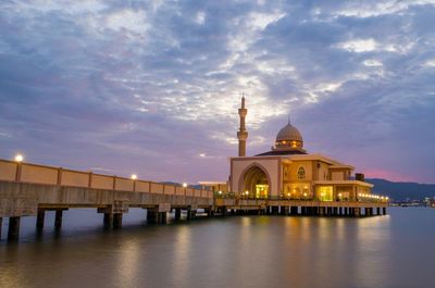 Illuminated mosque over sea against cloudy sky during sunset