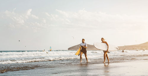 Couple holding surfboard walking on beach against sky