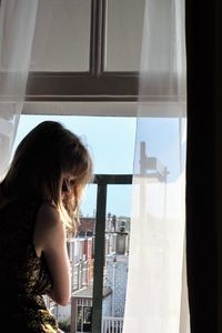 Side view of mid adult woman looking through window while standing at home