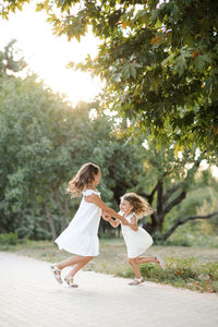 Two cute siblings girls wear similar white dress hoding hands and dance together in park
