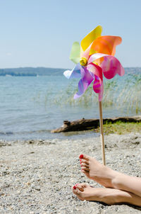 Low section of woman relaxing by pinwheel toy at beach against clear sky