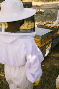 Unrecognizable crop beekeepers on protective costumes using bee smoker in apiary in summer