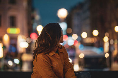 Young woman looking away while sitting in illuminated city at night