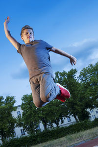 Teenage boy jumping in the air with arms raised on sports field against blue sky.