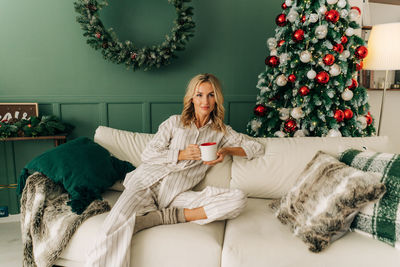A graceful young european woman in pajamas sits on a sofa on christmas eve and drinks coffee.