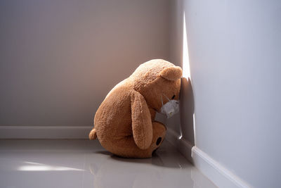 Close-up of stuffed toy bear with protective face mask kneeling on gray wall at home