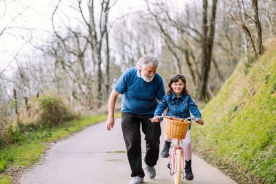 Distant caring grandfather helping active granddaughter riding bicycle with basket on road in countryside with green trees on summer day