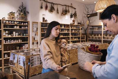 Young female with brown hair in casual clothe standing with small chihuahua dog in hand in front of woman seller and paying with smartphone in eco friendly shop