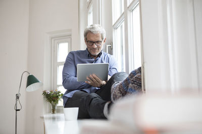 Mature man at home sitting at the window using tablet