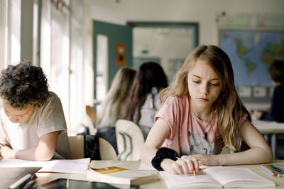 Female student studying from book while sitting by male friend in classroom
