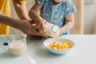 Close-up unrecognizable child and mother preparing corn flakes with milk for breakfast.