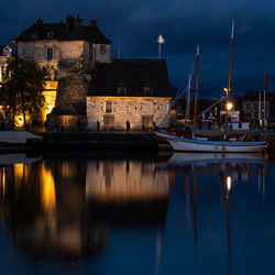 Sailboats moored on lake by illuminated buildings against sky at dusk