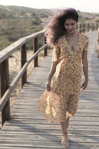 Slim delicate ethnic female with chest tattoo in light summer dress standing on wooden walkway and looking down while enjoying sundown