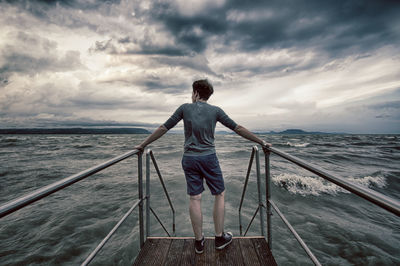 Rear view of man standing on pier in sea against cloudy sky