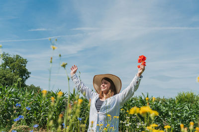Young woman with arms raised standing amidst flowering plants against sky