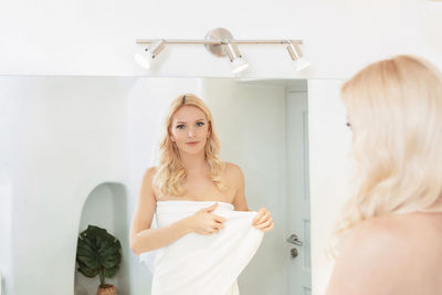 Blond woman in a white bathroom looking at her reflection in the mirror, wearing a towel