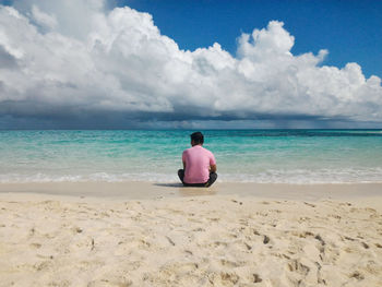 Rear view of mid adult man sitting at beach against cloudy sky during sunny day