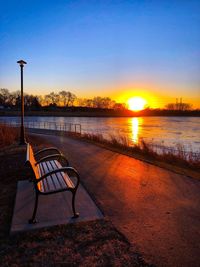Empty bench by lake against sky during sunset
