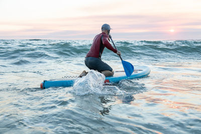 Back view of unrecognizable male surfer in wetsuit and hat on paddle board surfing on seashore