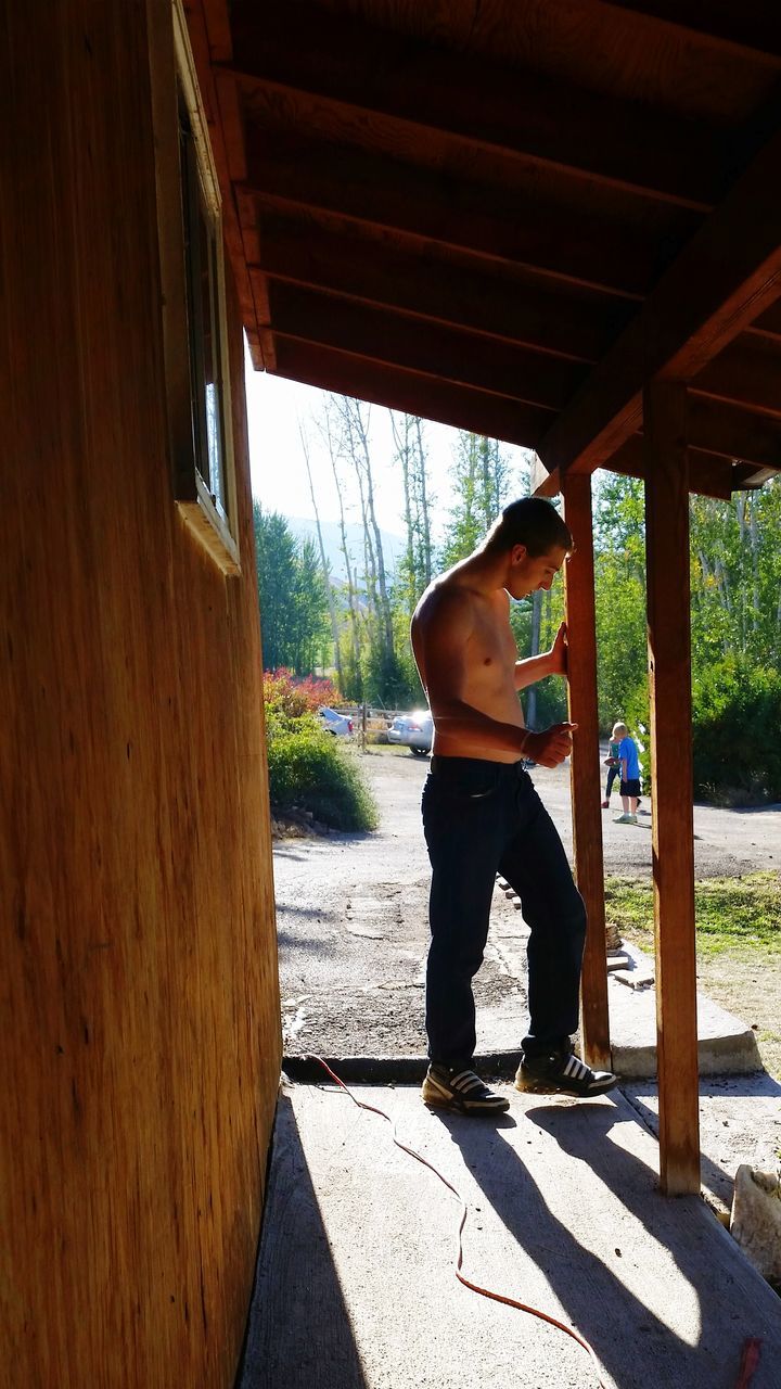 one person, full length, adult, architecture, day, sunlight, lifestyles, nature, leisure activity, wood, built structure, men, standing, shadow, casual clothing, young adult, tree, outdoors, footwear, side view, women, travel, house, relaxation, person