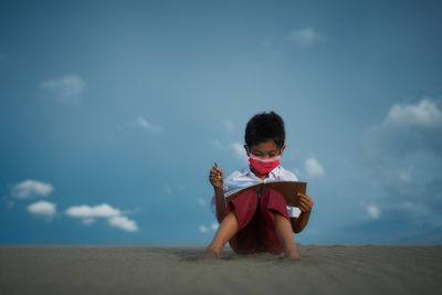 Little boy sitting and reading book at the beach under blue sky