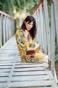 Young woman reading book while sitting on footbridge